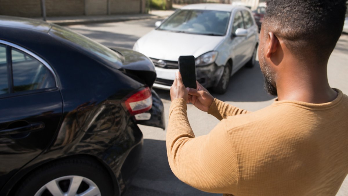 Driver photographing car damage at an accident scene where both drivers at fault may be determined.